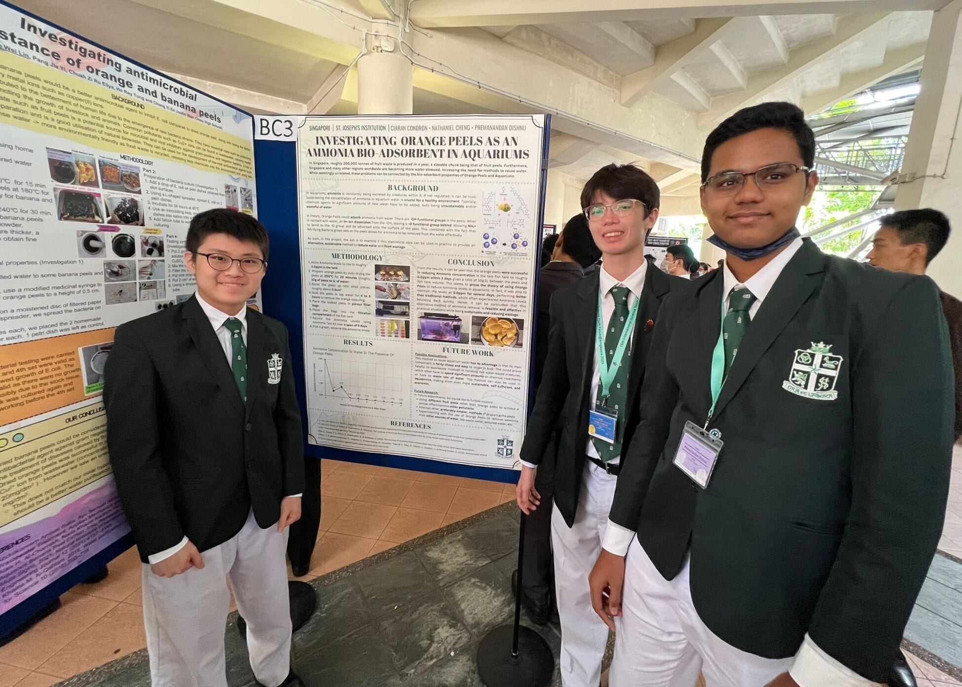 Three students in green blazers stand beside a science fair poster about orange peels as an ammonia bio-adsorbent.